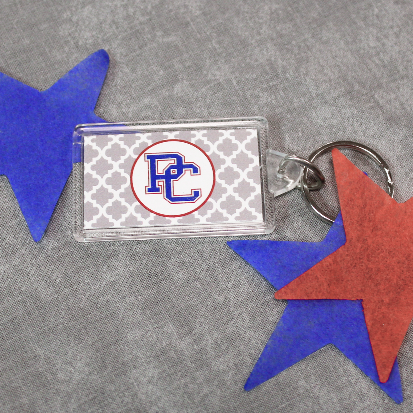 Picture of a rectangular acrylic keychain that has a grey and white diamond pattern background and then a white circle with the color block PC logo on it. In the product photo, there are red and blue stars surrounding the acrylic keychain.
