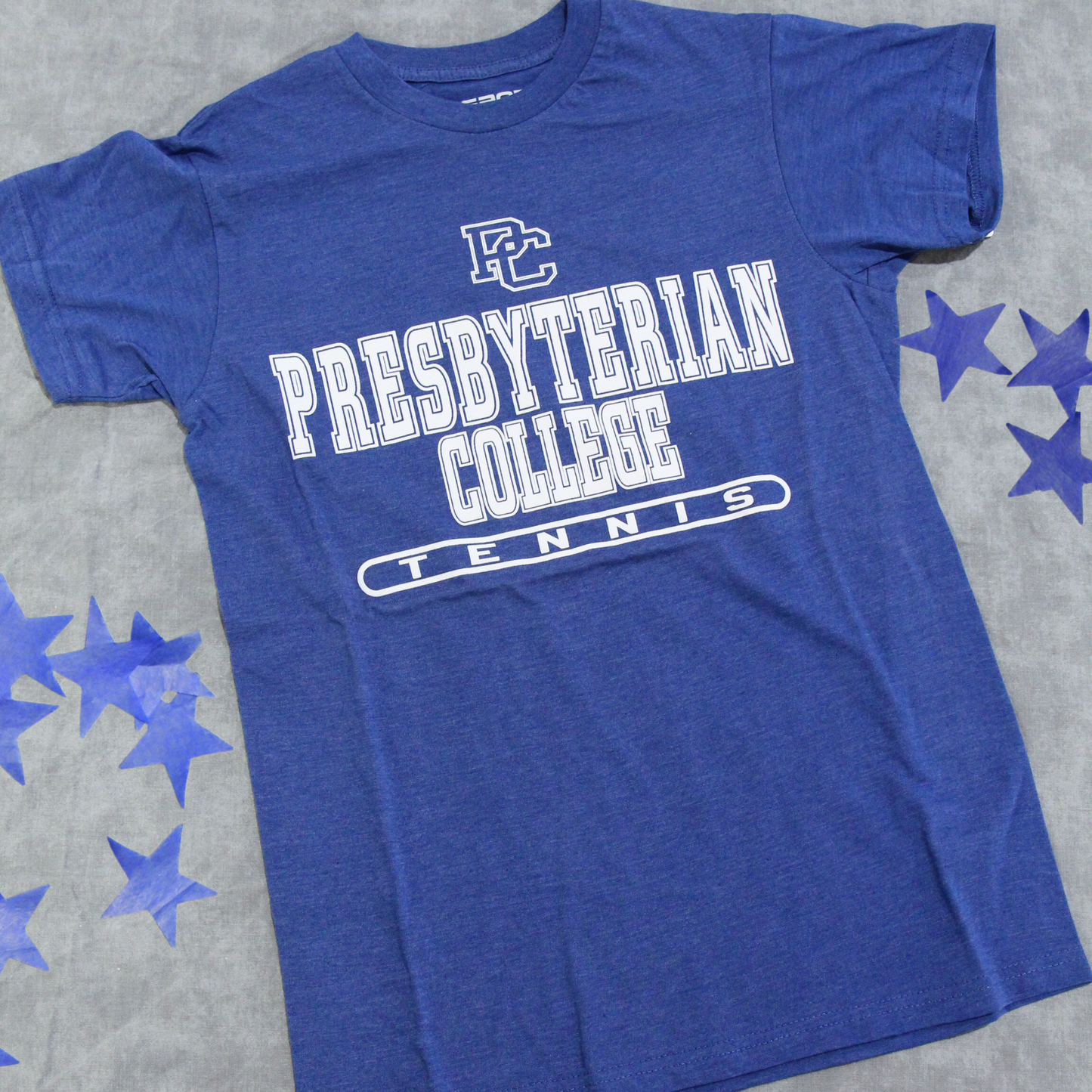 Product photo of a royal blue heather t-shirt with a white screen print graphic that has the block PC logo and then "Presbyterian College Tennis" on it. The product photo has blue stars in the background.