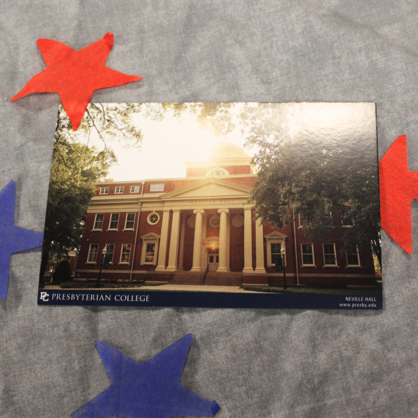 Product photo of the Neville Hall postcard. he postcard features a picture of Neville Hall during golden hour and framed by green trees. The background of the product phot has red and blue stars surrounding the postcard.
