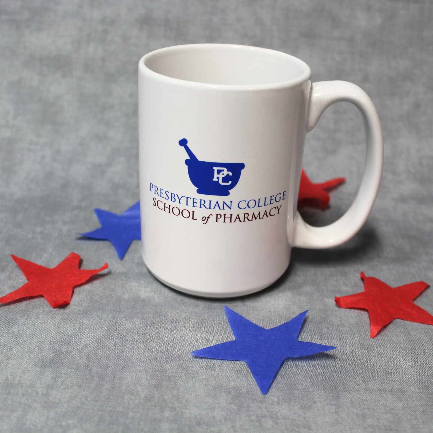 Product photo of a white mug with the Presbyterian College School of Pharmacy logo across the middle of the mug in blue and red ink. The background of the product phot has red and blue stars.