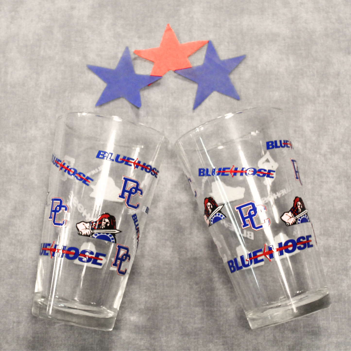 Another view of the two drinking glasses that feature the PC logo, Scotty the Scotsman Athletic logo, and the Blue Hose wordmark logo. Background features blue and red stars.