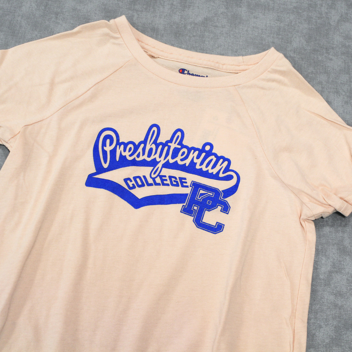 Up Close picture of the graphic on the light pink raglan sleeve youth champion shirt. The design features 'Presbyterian' in a cursive font similar to a baseball shirt with "college" underneath it and a block PC logo. The whole graphic is printed in royal blue.