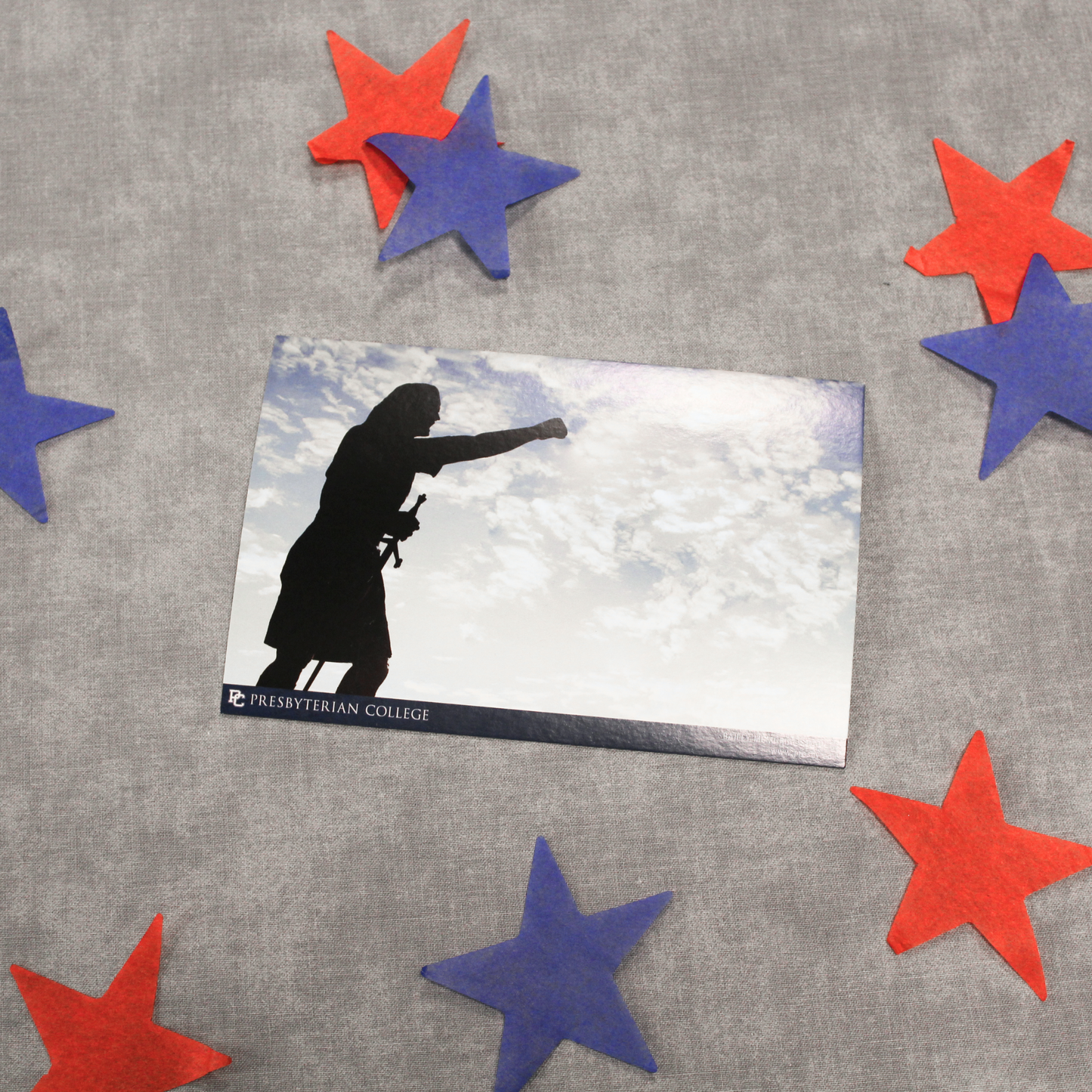 Postcard that features the silhouette of the statue of Cyrus the Scotsman from in front of Bailey Stadium against a background of a cloudy sky. The background of the picture of the postcard features red and blue stars.