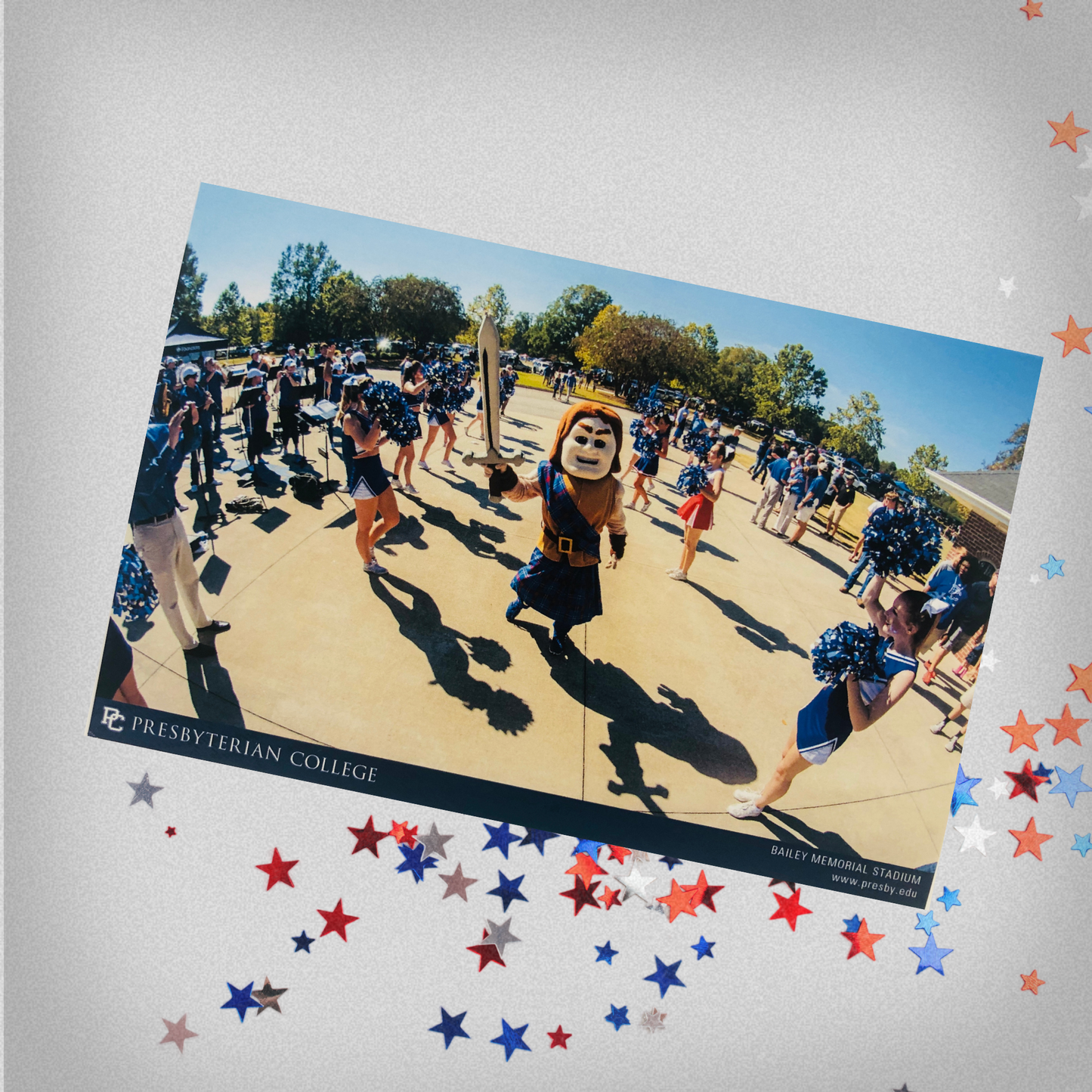 Picture of the Bailey Stadium Cheer Postcard that features Scotty the Scotsman Mascot carrying a sword surrounded by the Presbyterian College Cheer team and fans out front of the football stadium. The background of the product photo features red, white, and blue stars.