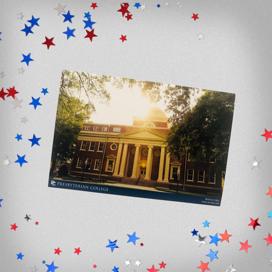 Product photo of the Neville Hall postcard. he postcard features a picture of Neville Hall during golden hour and framed by green trees. The background of the product phot has red, white, and blue stars surrounding the postcard.
