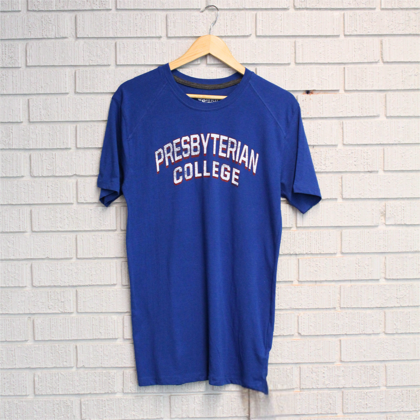 Photo of a royal blue fit flex t-shirt with a distressed screen print graphic in white and red ink of "Presbyterian College" against a white brick backdrop