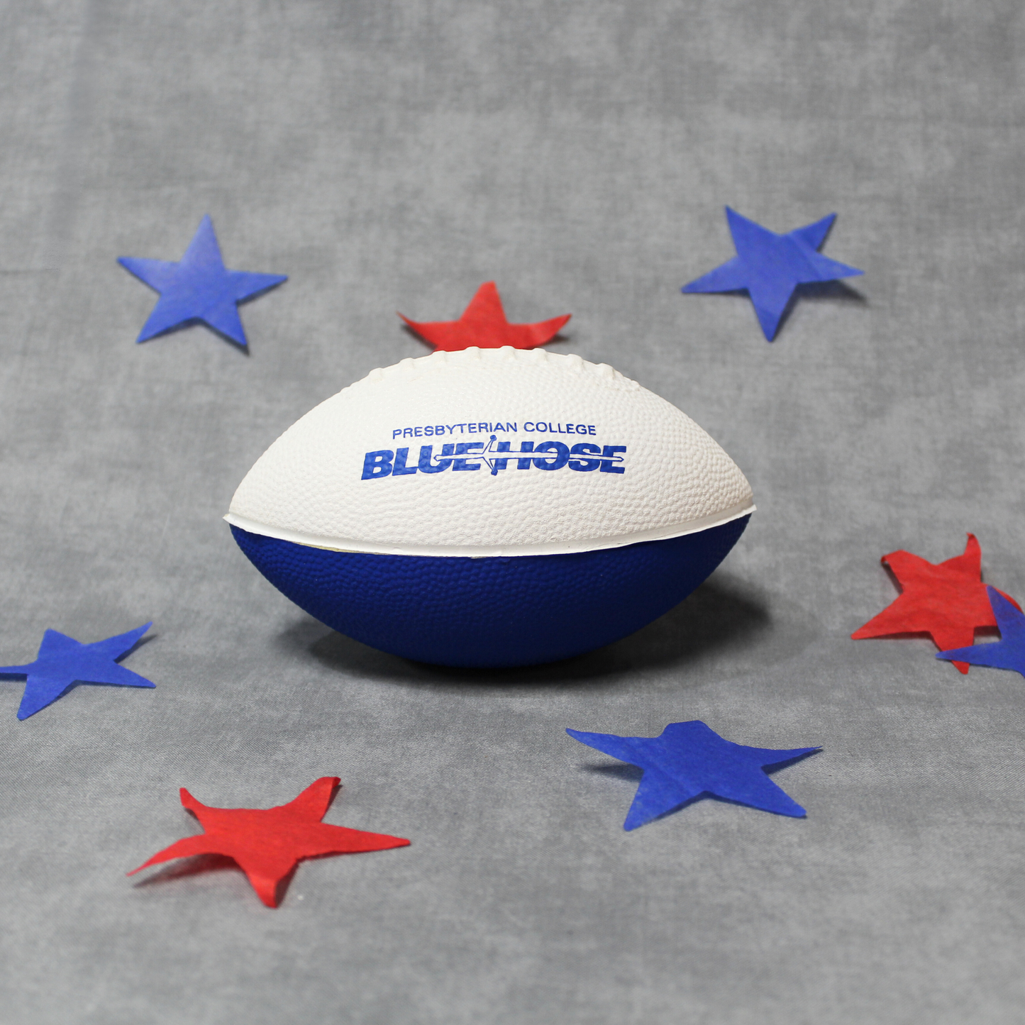Photo of a small white and royal blue foam football with the Presbyterian College Blue Hose athletic wordmark printed across the top of the football in royal blue ink. The backdrop has red and blue stars surrounding the football.