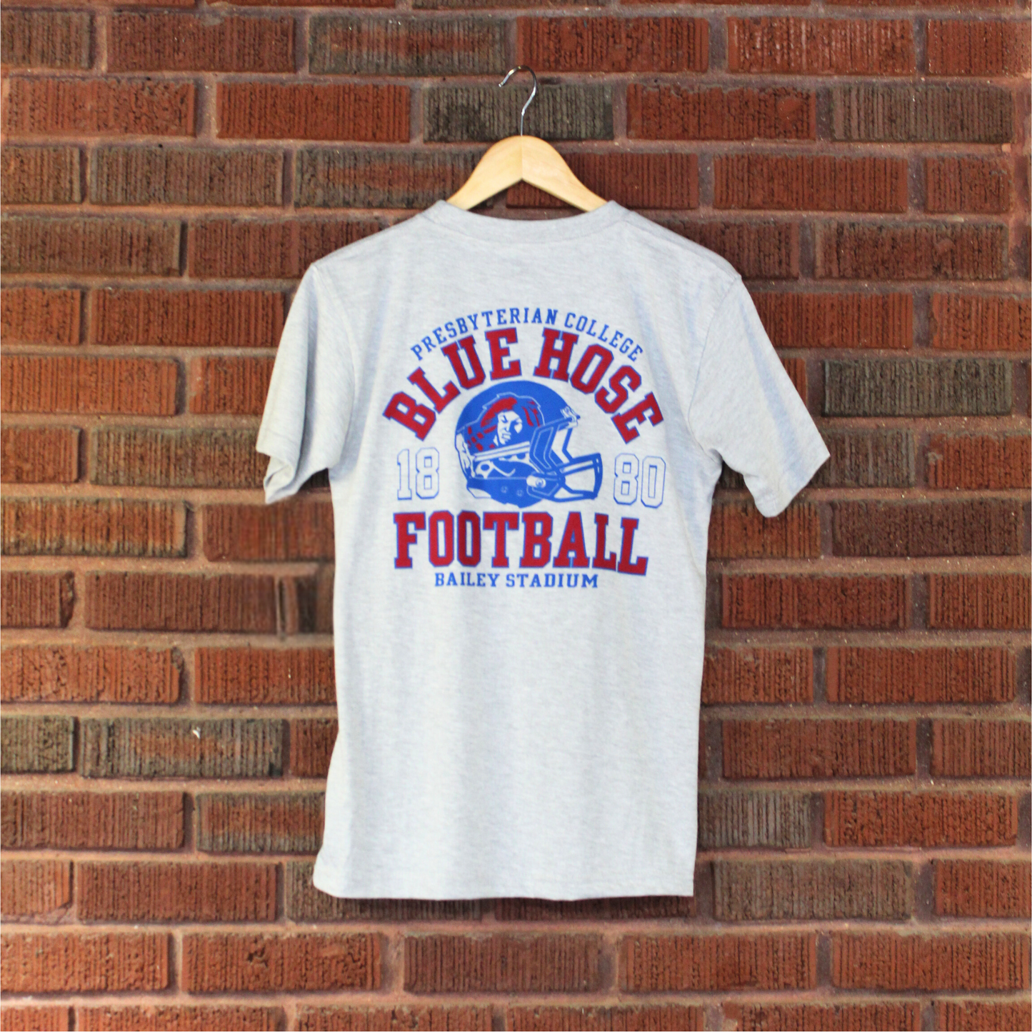 Product photo of the back of a light grey t-shirt that features a red and blue screen print of a football helmet with scotty the scotsman on it, along with the words Presbyterian College Blue Hose 1880 Football Bailey Stadium on it.