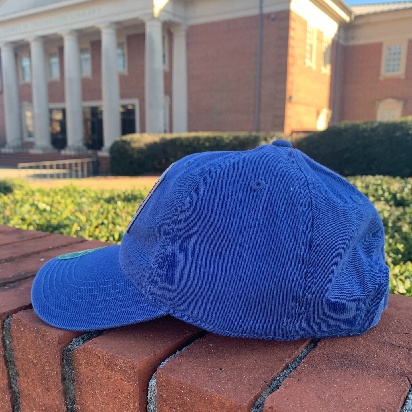 side view of a royal blue hat with a red and blue fabric patch on the front that has the PC logo on it, "1880", and "Presbyterian College". The hat is sitting on a brick wall in front of the Thomason Library.