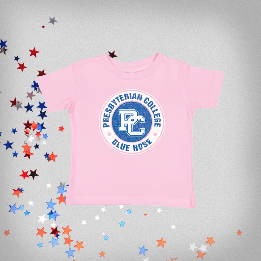 Product photo of a pink toddler t-shirt with a distressed blue and white Presbyterian College design, the background of this photo has red white and blue stars on it.