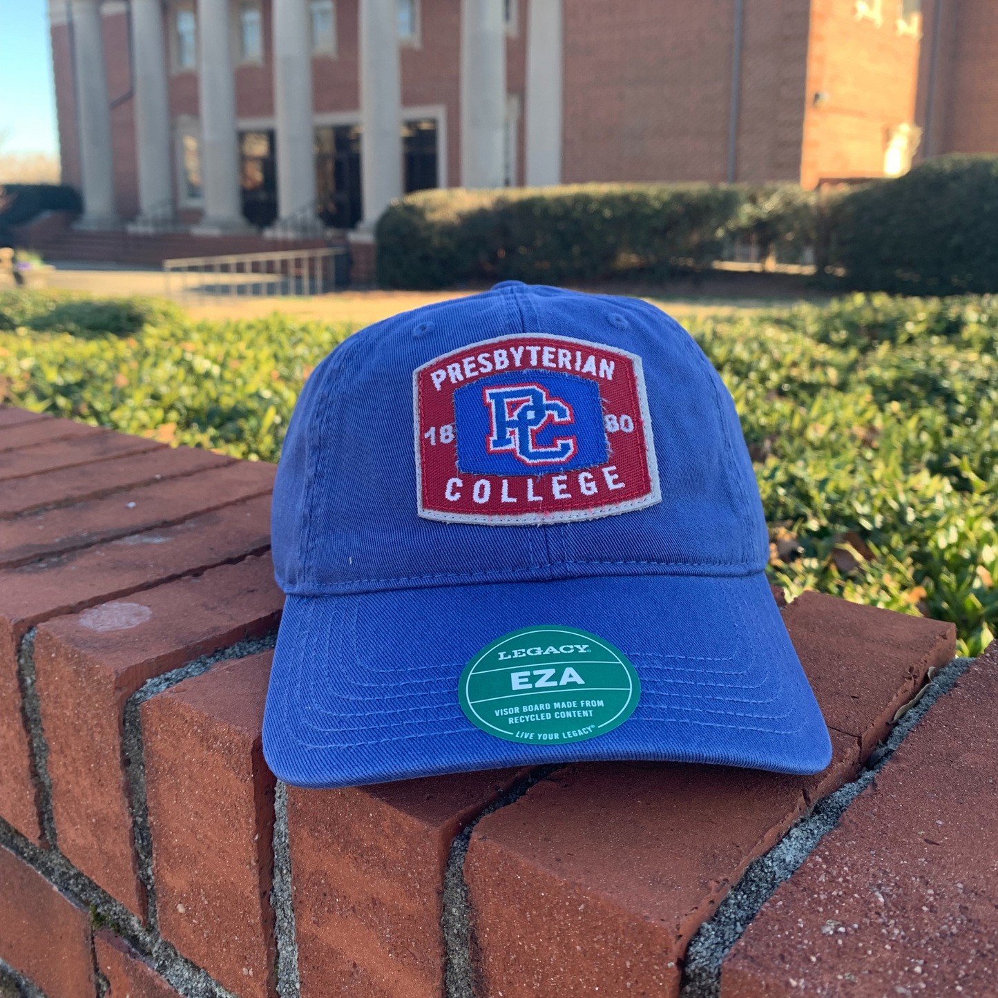 Product photo of a royal blue hat with a red and blue fabric patch on the front that has the PC logo on it, "1880", and "Presbyterian College". The hat is sitting on a brick wall in front of the Thomason Library.