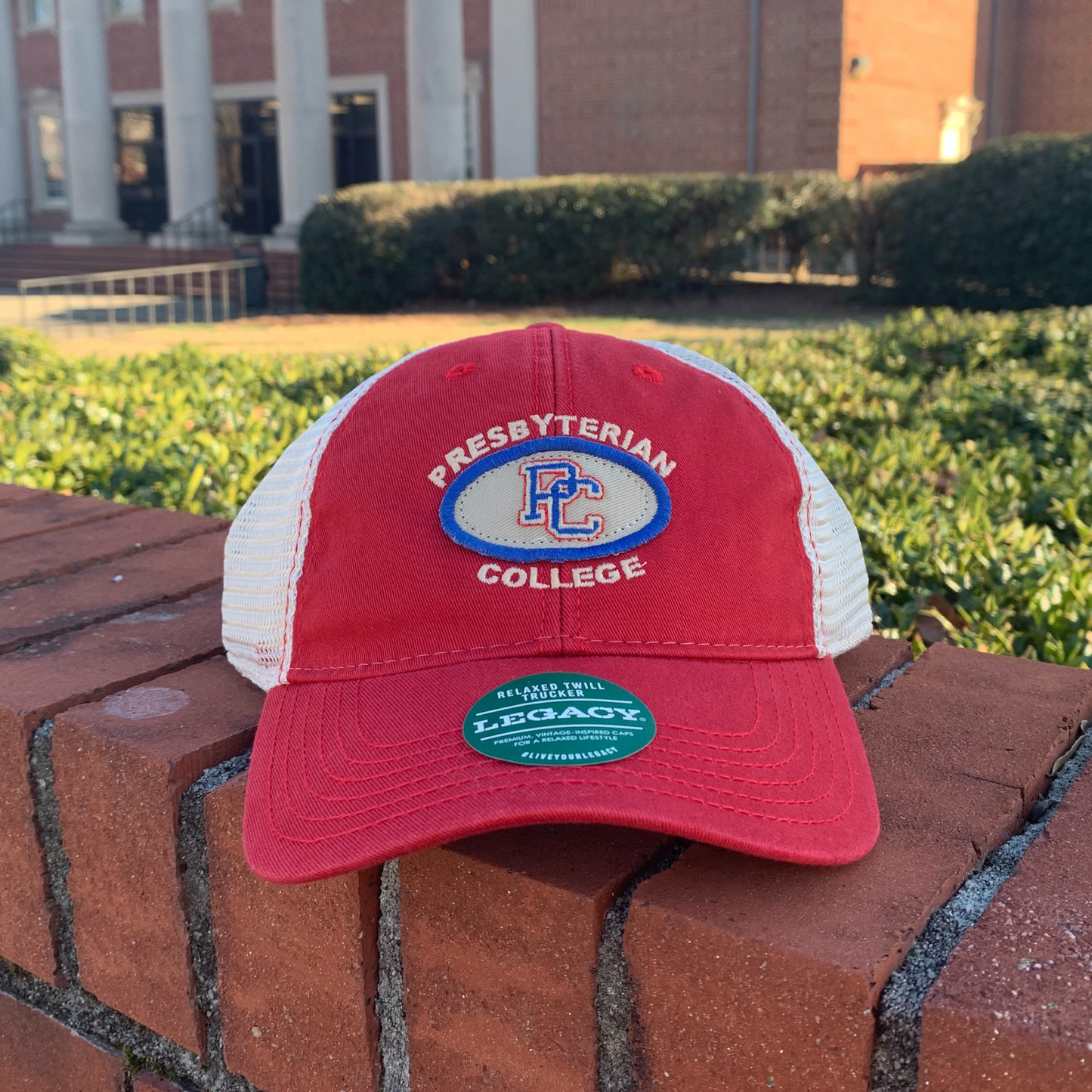 Product photo of a red trucker hat with a grey and royal patch on the front with a color block PC embroidered on it and the words "Presbyterian College" embroidered in white around the patch.