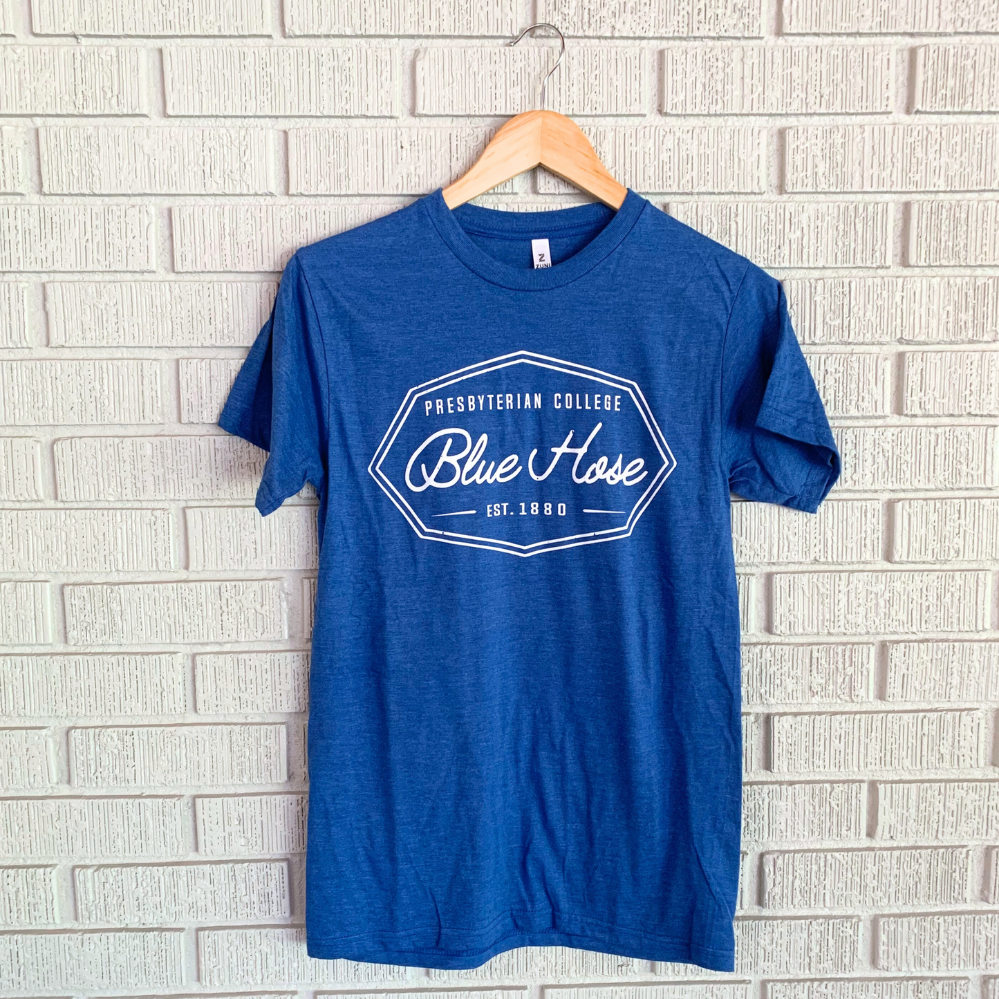 A blue t-shirt with 'Blue Hose' and 'Presbyterian College' text logo on the front, displayed on a wooden hanger against a brick wall background.