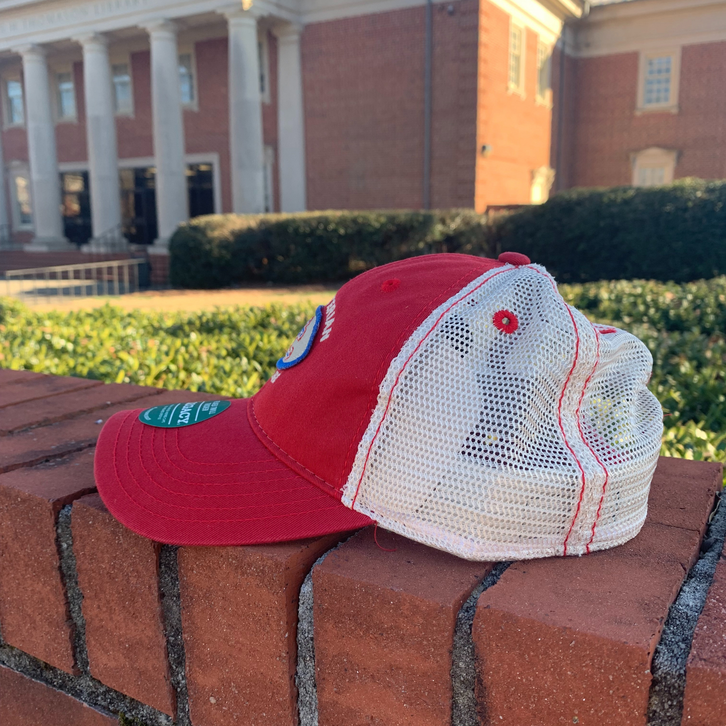Product photo of the side view of a red trucker hat with a grey and royal patch on the front with a color block PC embroidered on it and the words "Presbyterian College" embroidered in white around the patch.