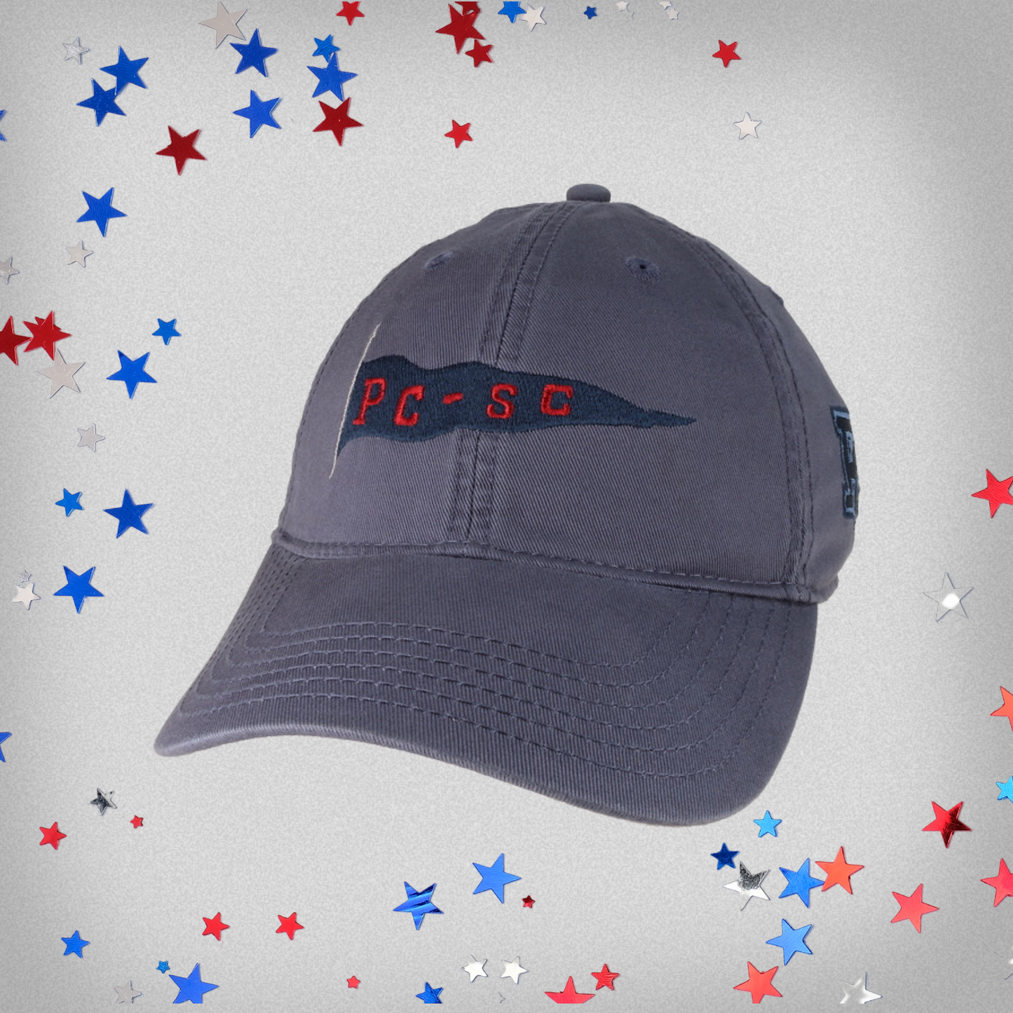 Slate Blue hat with an embroidered PC-SC pennant flag across the front of the hat, with a blue embroidered PC logo on the side. This product photo has red, white, and blue stars on the background.