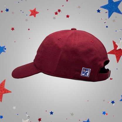 A maroon baseball cap with a small embroidered logo patch on the side, shown against a light gray background decorated with red, white, and blue glitter stars.